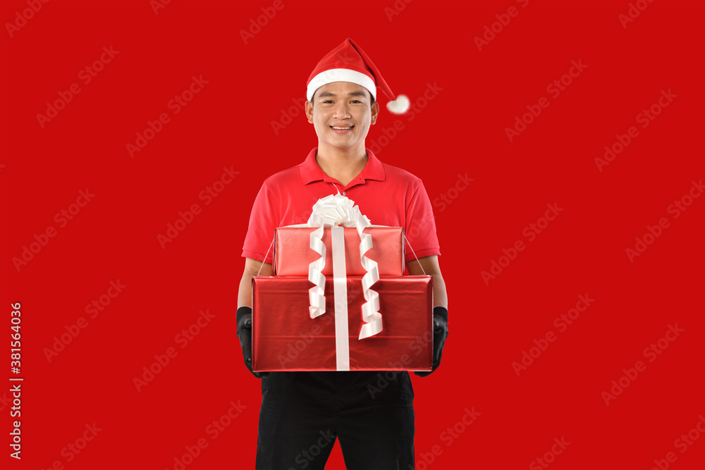 Happy young Asian delivery man in red uniform, christmas hat carry boxes of presents in hands on red