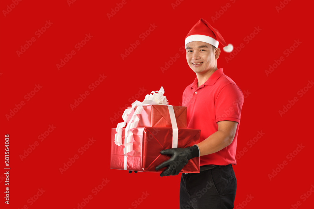 Happy young Asian delivery man in red uniform, christmas hat carry boxes of presents in hands on red