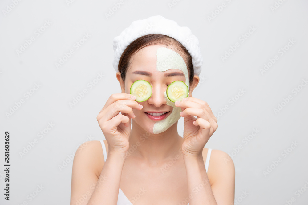 Portrait of young Asian woman with clay mask and holding cucumber piece against white background.