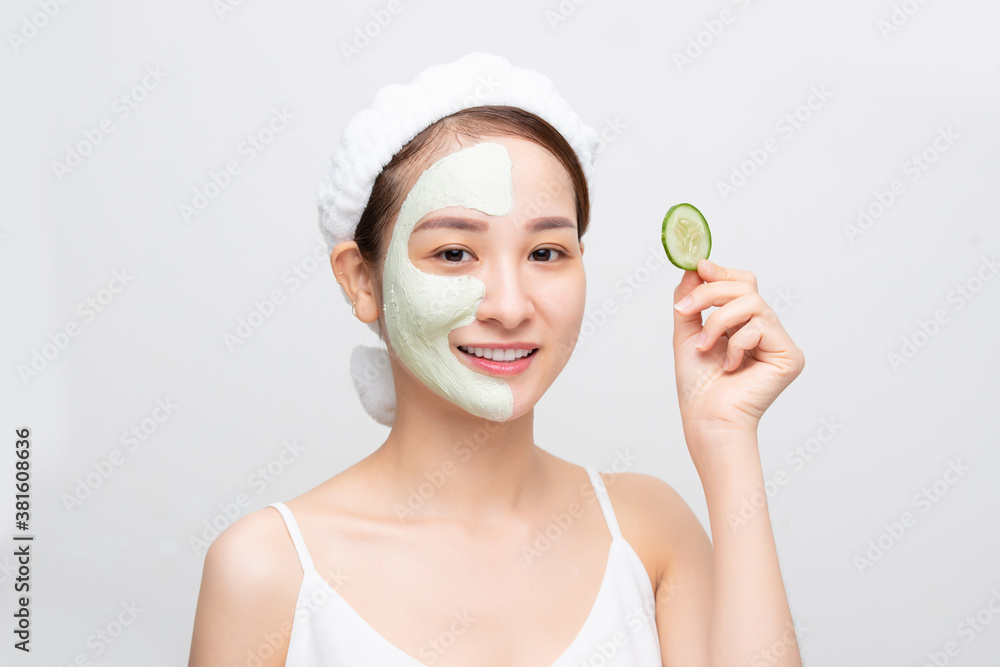 Beautiful young Asian woman with clay mask face holding cucumber piece over white background.