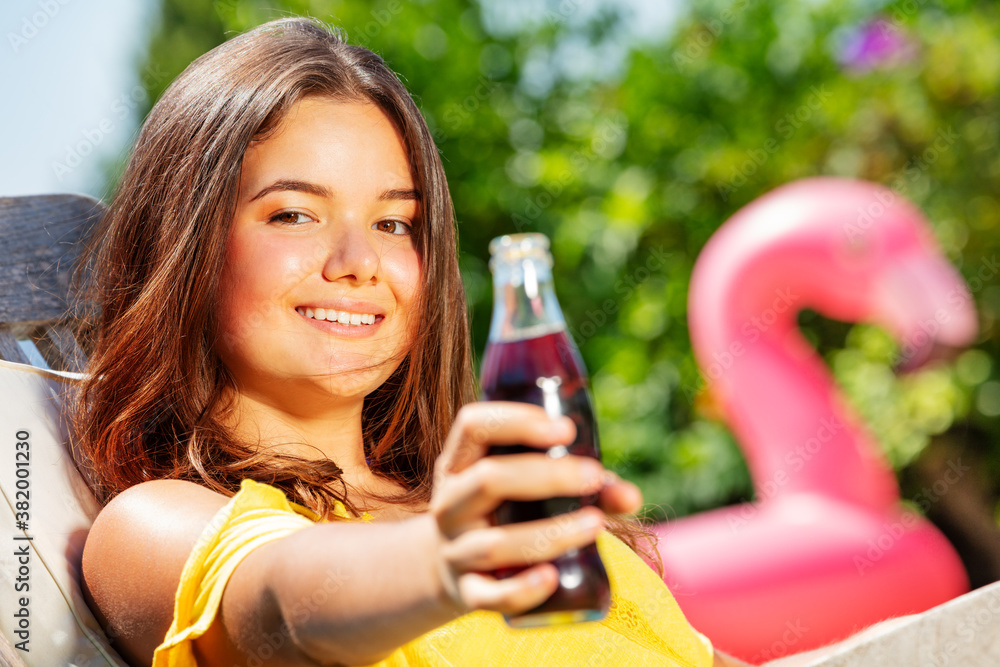 Happy teenage long hair girl give soda glass bottle sitting in the garden on chaise longue