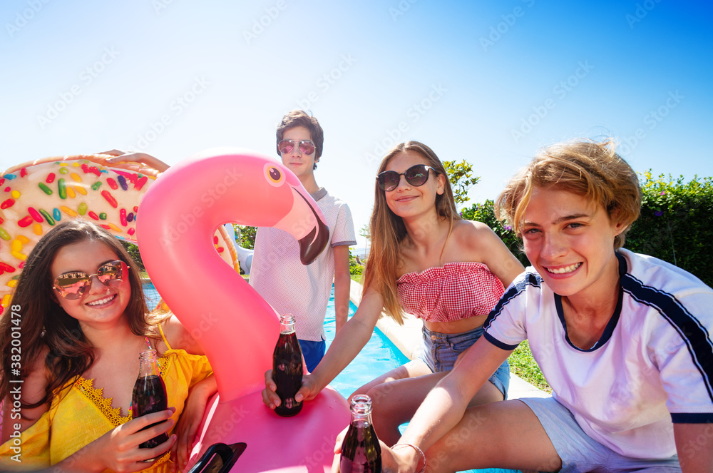 Teenage boys and girls chat, drink soda celebrating sitting on the border pool with inflatable buoys