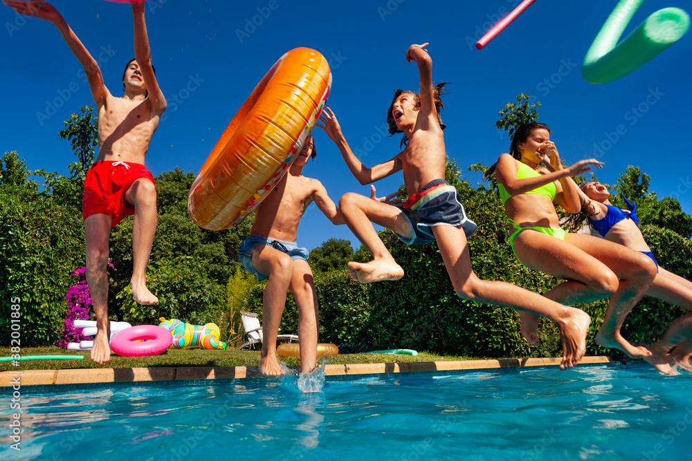 Group of happy teen kids jump and dive in water throwing inflatable toys in the air splashing into t