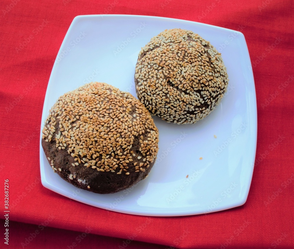 Homemade rye buns with sesame seeds on a white glossy plate. Against the background of red linen fab