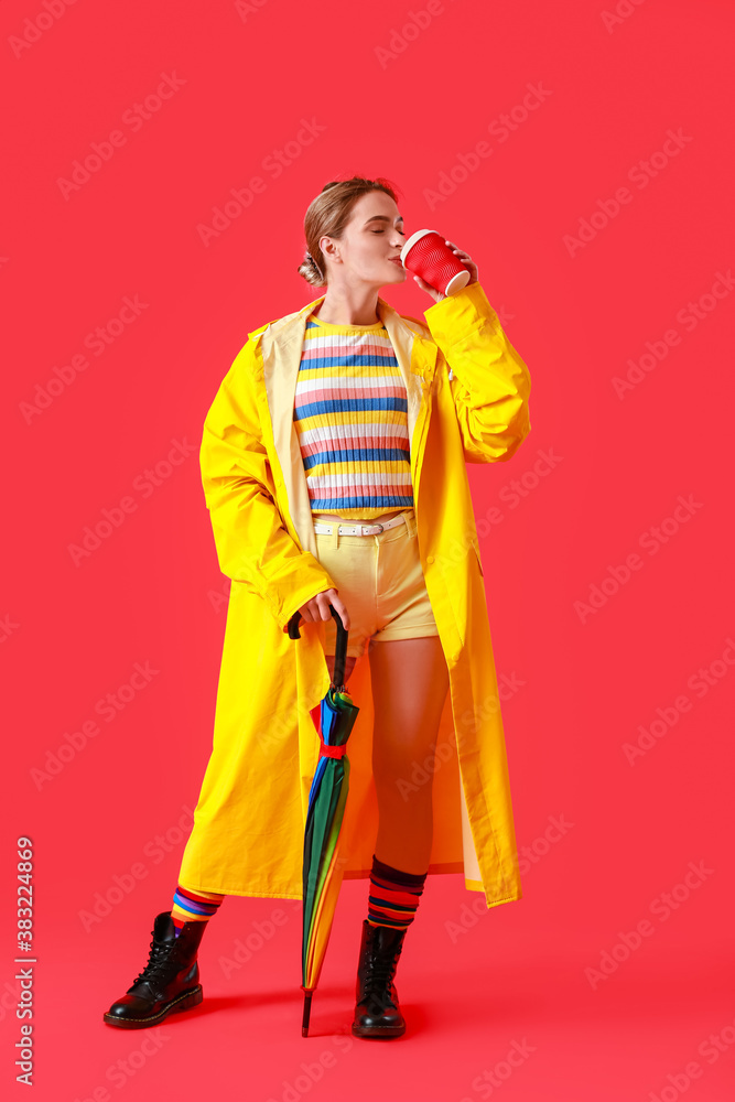 Beautiful young woman in raincoat, with coffee and umbrella on color background