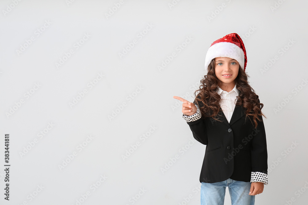 Cute little girl in Santa hat pointing at something on light background