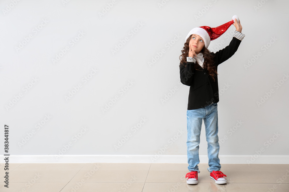 Thoughtful little girl in Santa hat near light wall