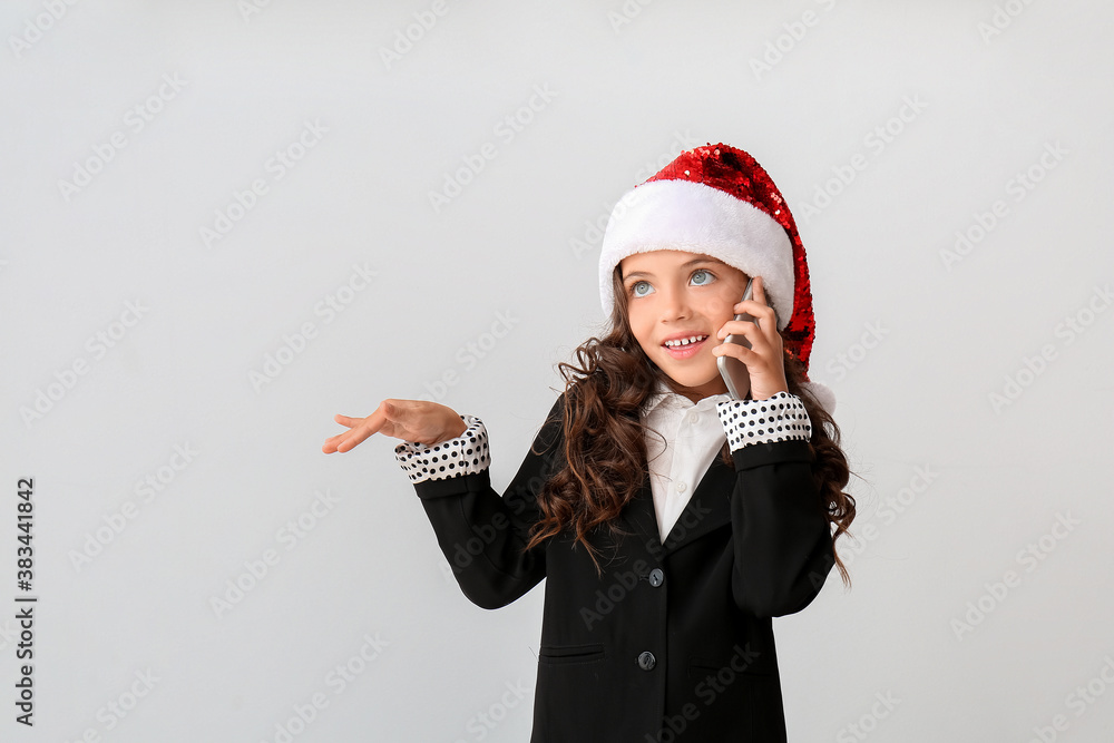 Cute little girl in Santa hat talking by phone on light background