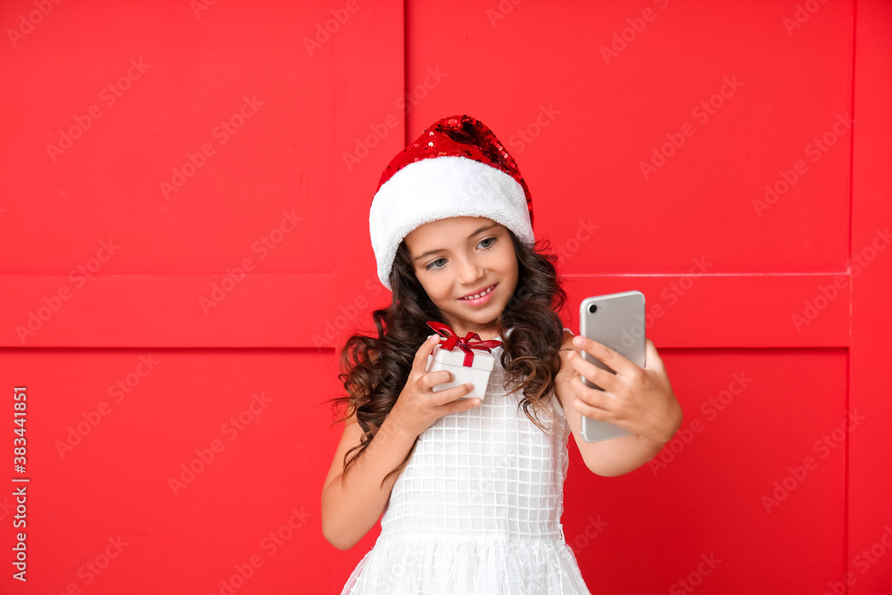 Cute little girl in Santa hat taking selfie on color background