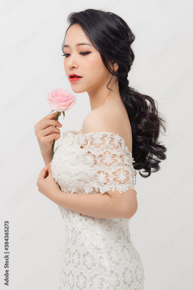 Cheerful young Asian lady wearing white dress and holding flower bouquet over white background.