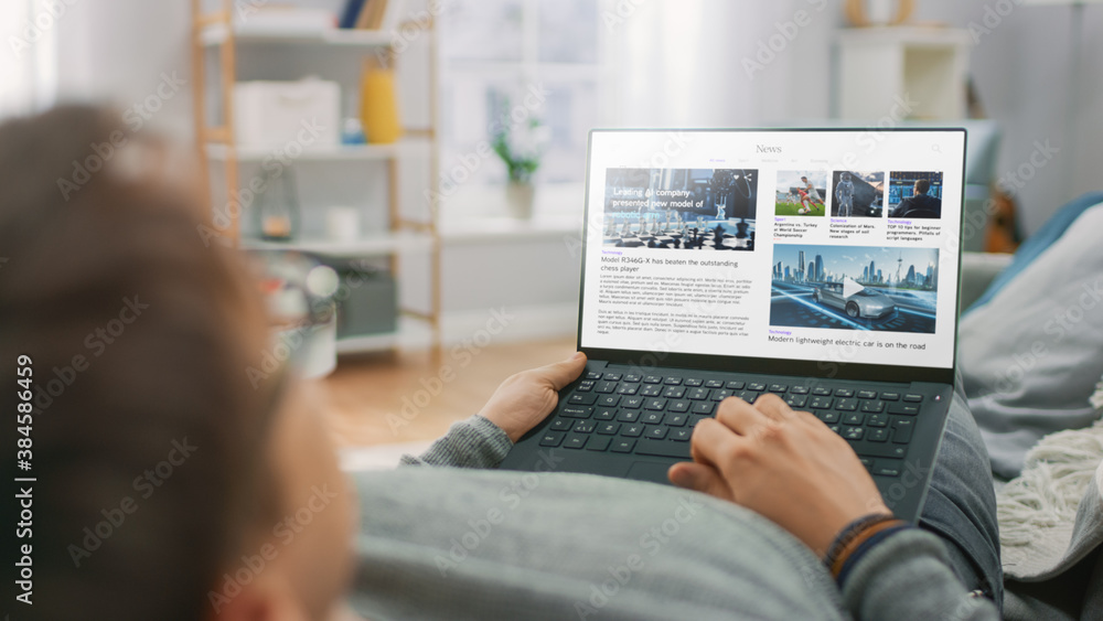 Young Man at Home Laying on a Couch and Using Laptop Computer for Reading News about Technological B