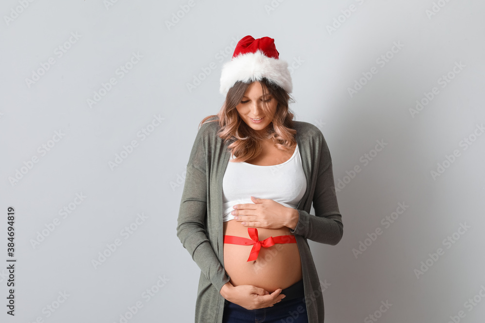 Beautiful pregnant woman with Santa Claus hat and ribbon around her waist on light background