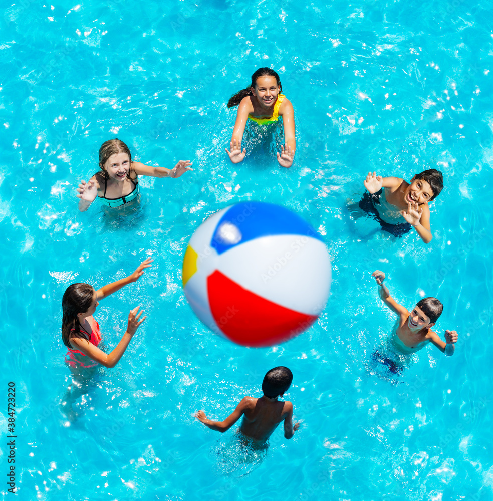 Group of little children in pool stand in circle lift hands up and play volleyball