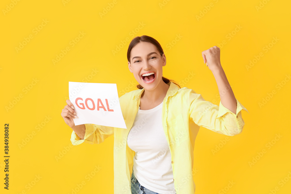 Happy young woman holding paper with word GOAL on color background