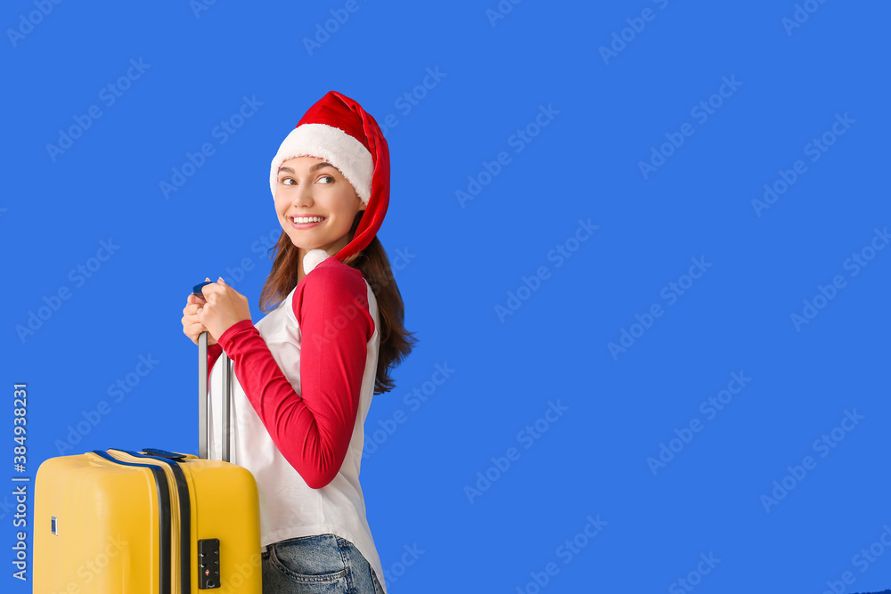 Beautiful young woman in Santa hat and with luggage on color background. Christmas holidays concept