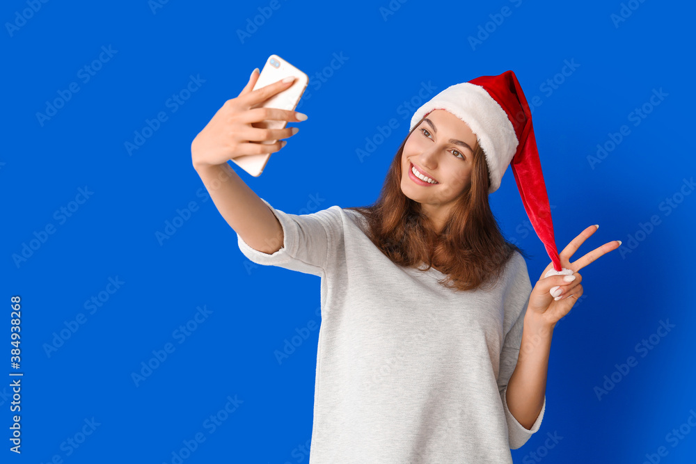 Beautiful young woman in Santa hat taking selfie on color background