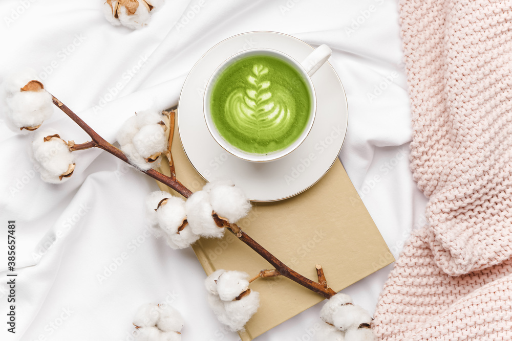 Composition with cup of matcha tea, beautiful cotton branch and book on bed