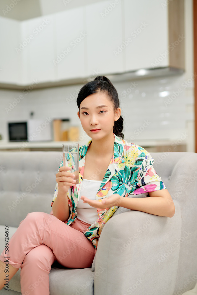 Happy woman taking vitamin pill holding glass sitting on a couch at home