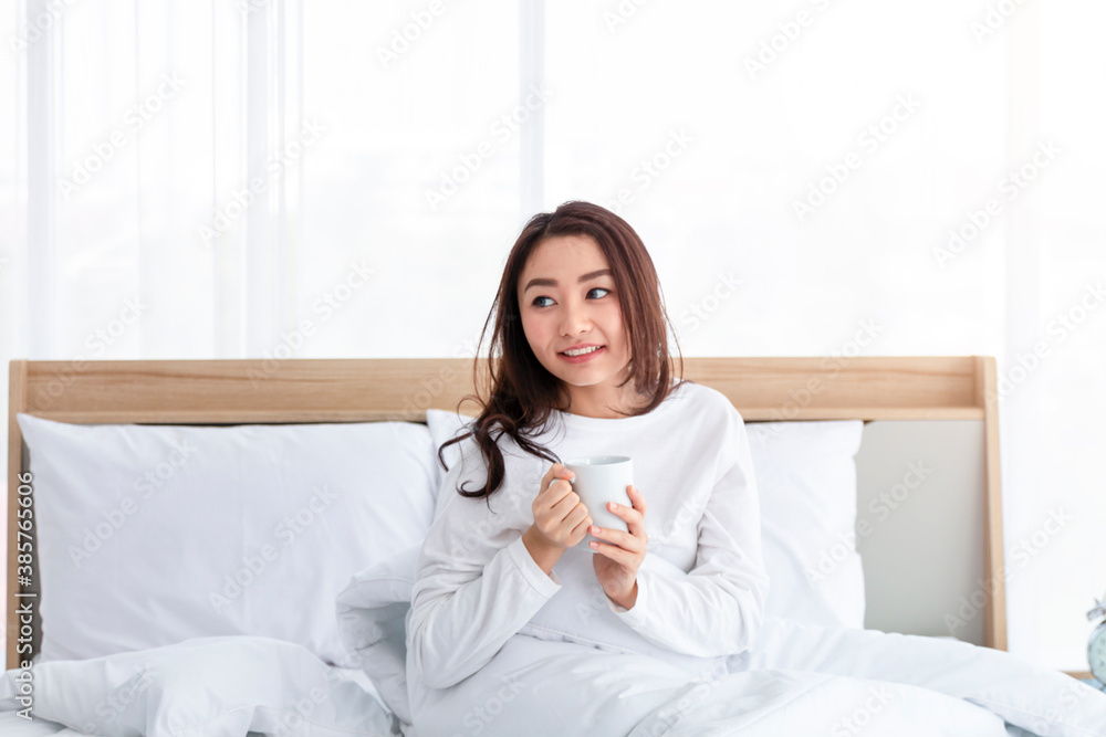 Young girl in white with coffee cup on bed in the morning. Portrait of beautiful young woman drinkin