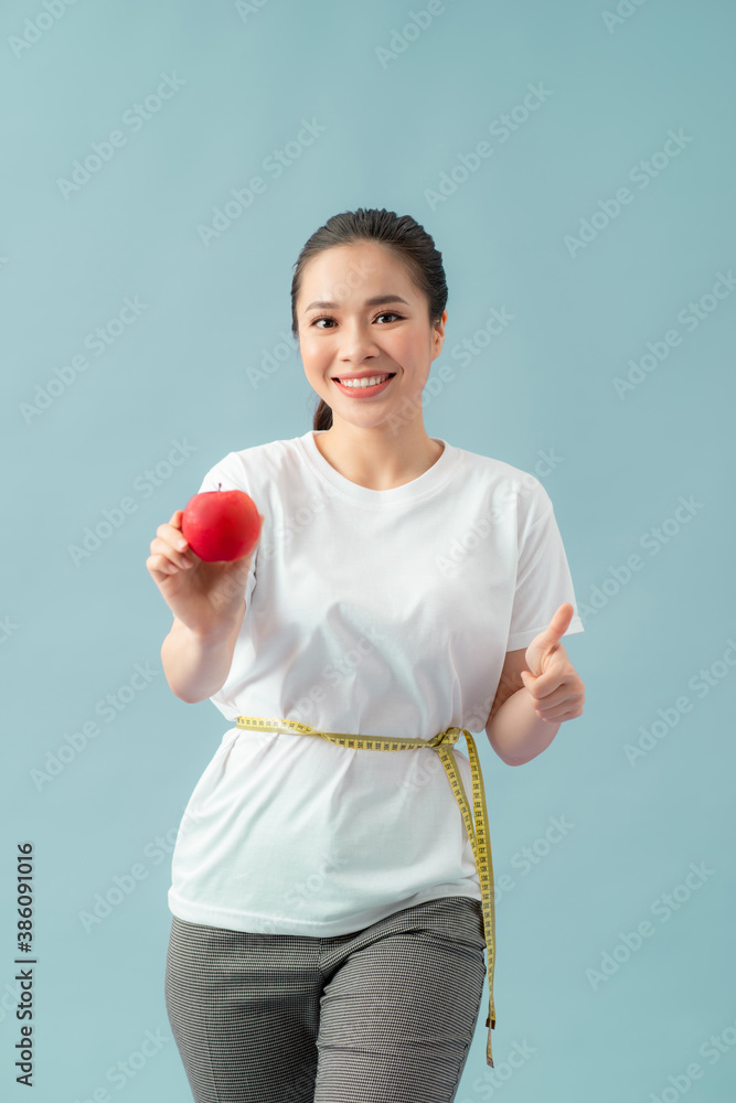 Beautiful sporty woman using tape measure eating apple fruit, showing thumb up, smiling happy with o