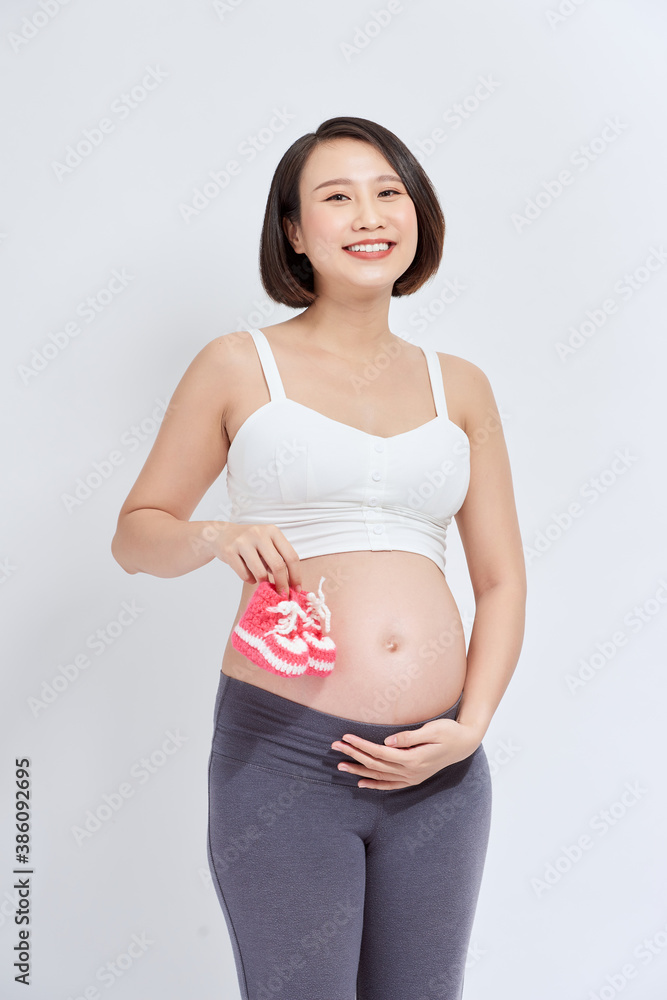 dreamy pregnant woman holding baby booties near tummy on white