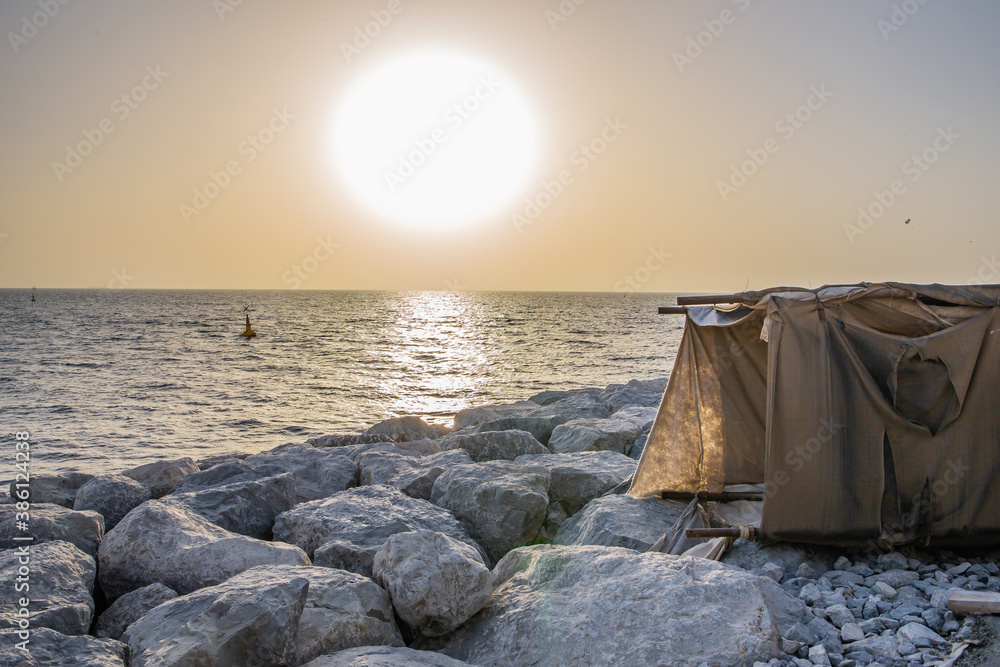 Temporary fabric tent made of old rugs standing on a stone jetty, with sunset in the background.