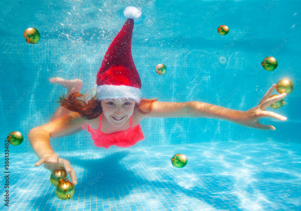 Happy girl dive and swim underwater with Christmas decoration balls wearing Santa Claus hat in the p