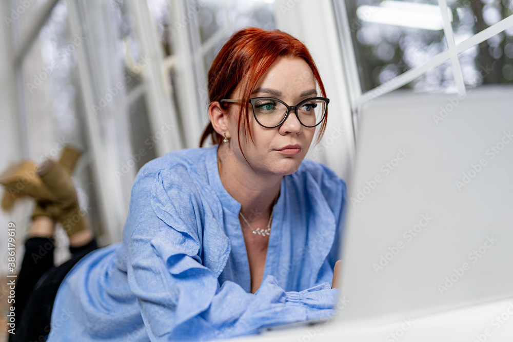 Young woman laying on floor using laptop. Big panoramic windows on the blured background. Working fr