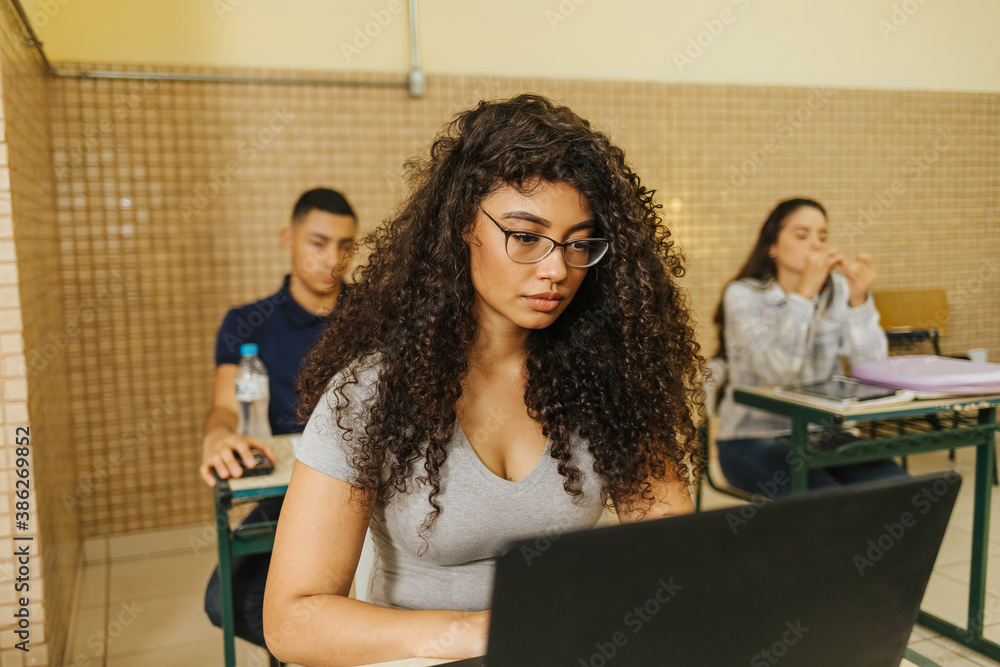 curly haired latin woman student writing on computer in a classroom. woman wearing glasses