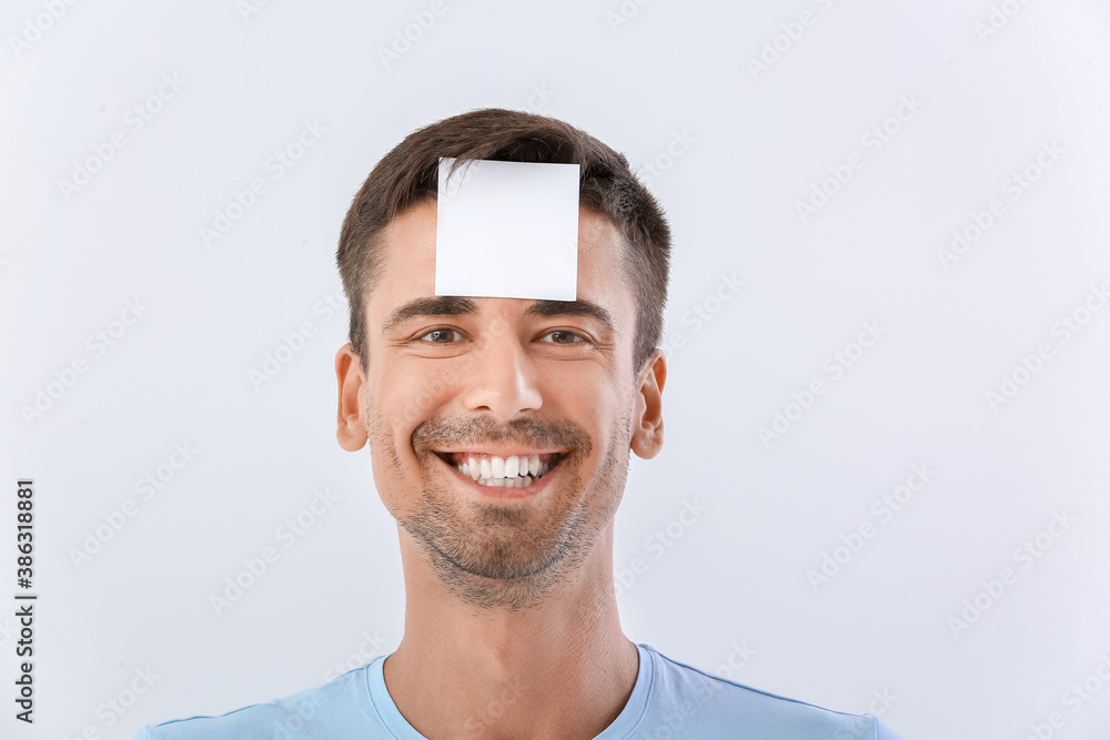 Young man with small blank paper sheet on his forehead against white background