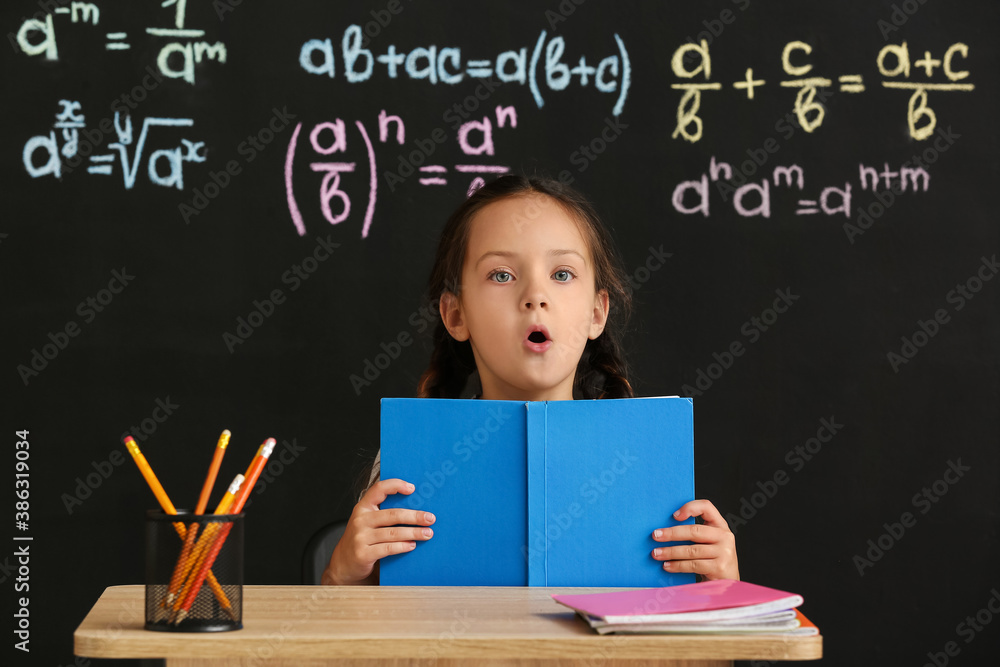 Cute pupil with book sitting at desk during lesson in classroom