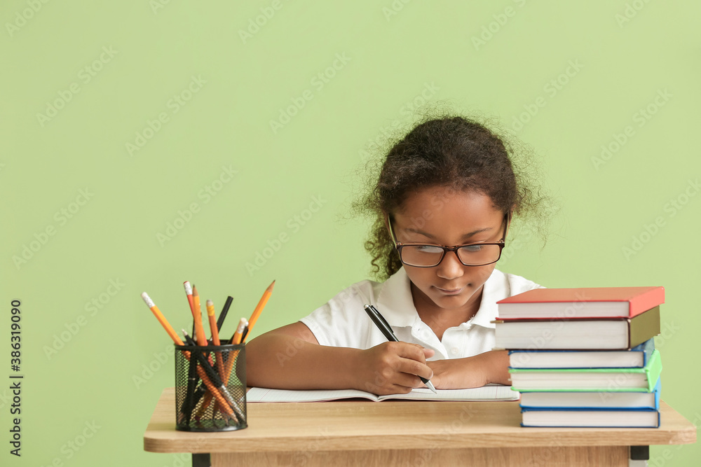 Cute African-American schoolgirl sitting at desk against color background