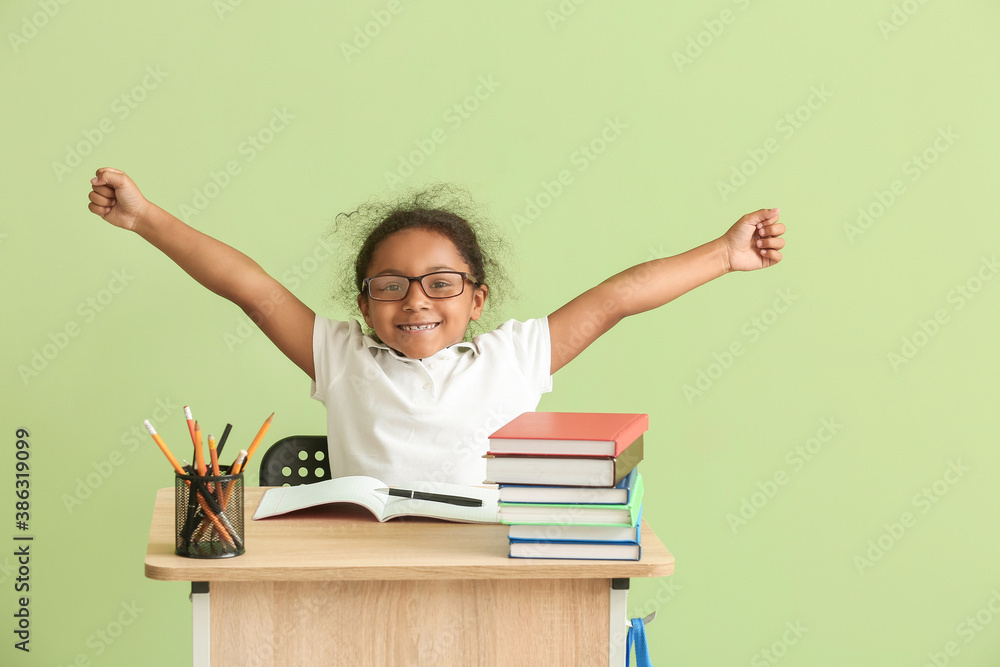 Happy African-American schoolgirl sitting at desk against color background