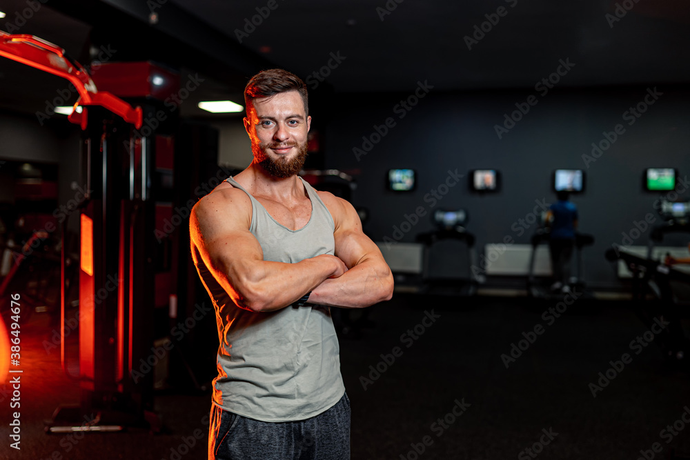 Full length of smiling sportive young man standing crosshands. Dark background of a modern gym.