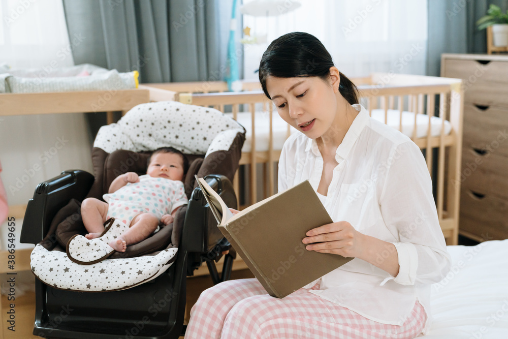 亚洲新妈妈坐在她年幼的孩子身边，正在给她的新生儿读故事绘本。nurturin