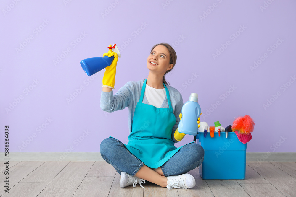 Young woman with cleaning supplies sitting near color wall