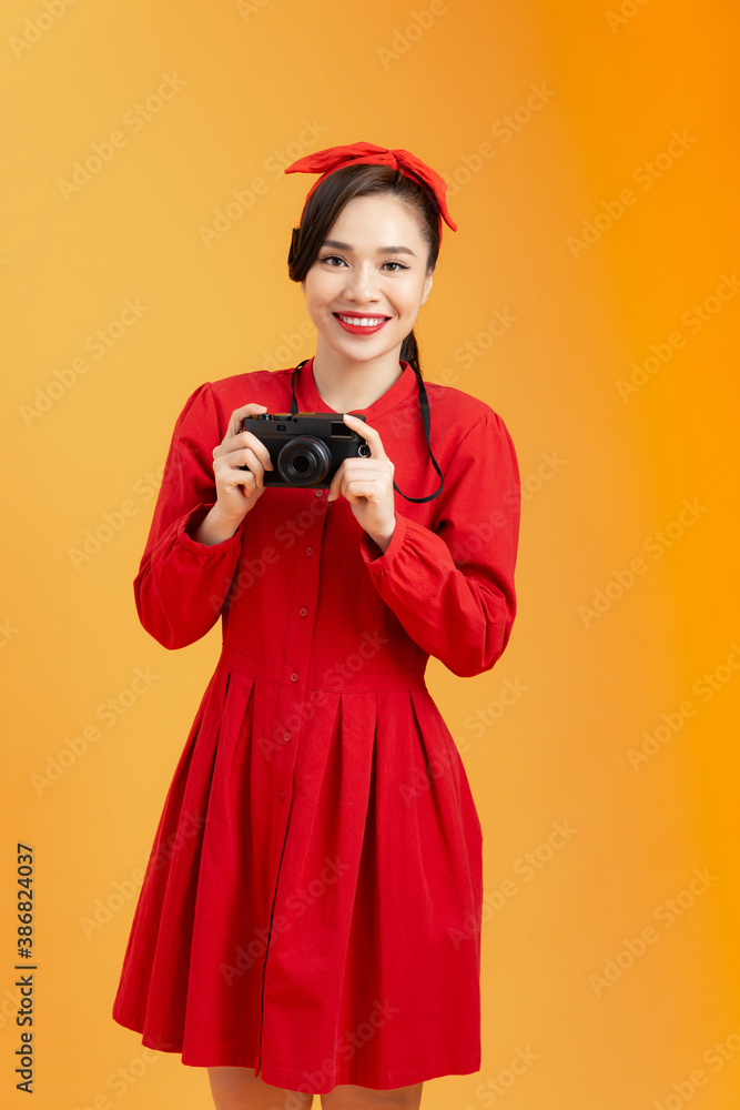 Attractive young pretty Asian woman in red dress holding camera over orange background.