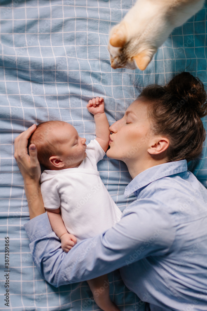 Mother lies in bed with newborn son. Mom kisses the baby. The pet is a family member. Cat is an anim