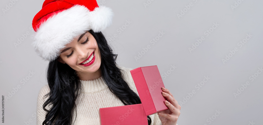 Young woman with santa hat opening a Christmas gift box on a gray background