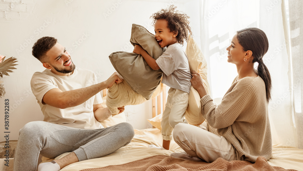 happy multiethnic family mom, dad and child  laughing, playing, fights pillows and jumping in bed   