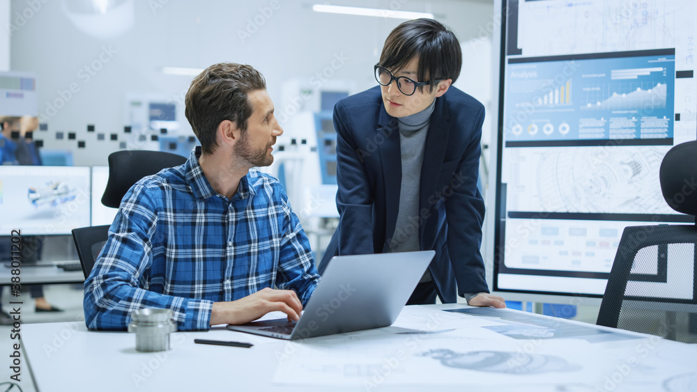 Modern Factory: Industrial Engineer Sitting at His Desk, Working on Laptop Computer, Talks with Chie