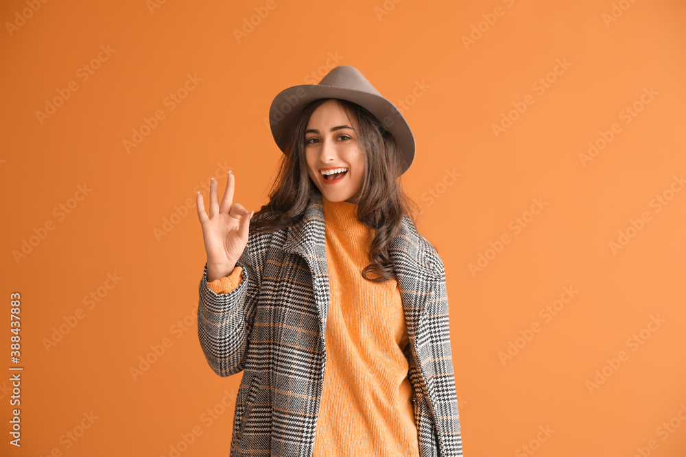 Stylish young woman in autumn clothes showing OK gesture on color background