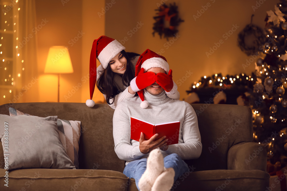 Young woman covering eyes of her husband who is reading book at home on Christmas eve