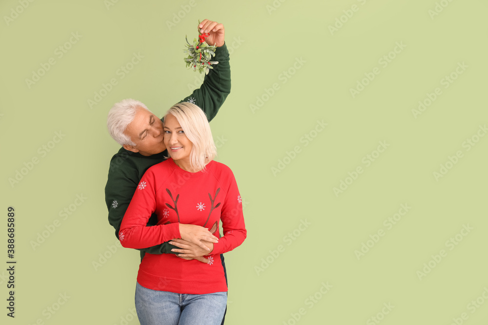 Mature man kissing his wife under mistletoe branch on color background