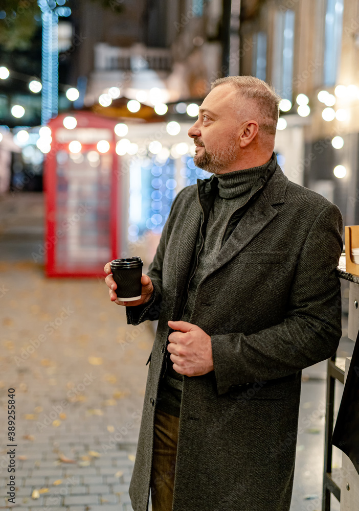 Coffee break. Mature man in elegant coat smiling nicely while having coffee. Evening street.