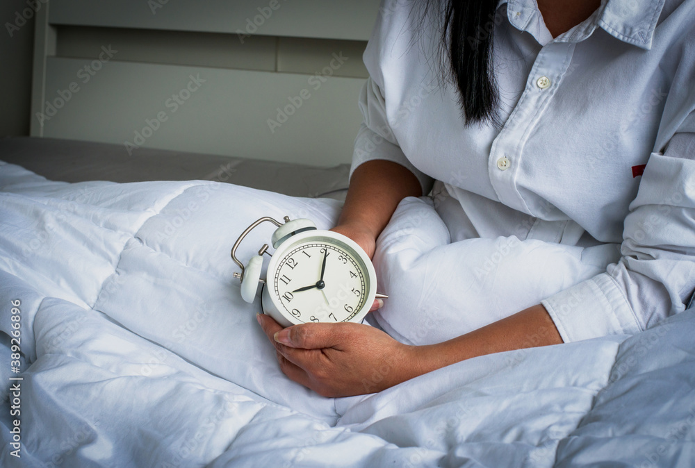 a young Asian woman sitting in bed holding the white alarm clock to set the alarm for the day after,