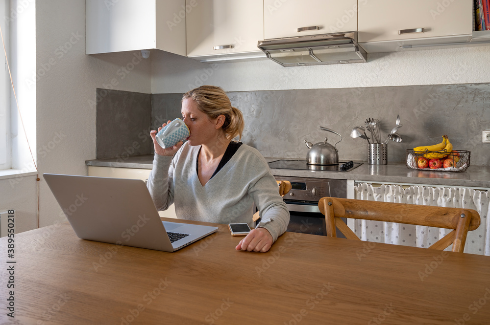 Femme dans sa cuisine en télétravail devant son ordinateur buvant un café