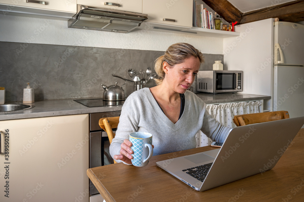 Femme dans sa cuisine en télétravail devant son ordinateur buvant un café