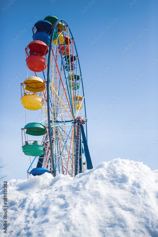 View of a large snowdrift and a Ferris wheel with colorful cabins in an amusement park. Vintage ride