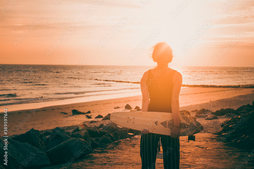 A girl holding her longboard on the sandy beach at sunset in summer